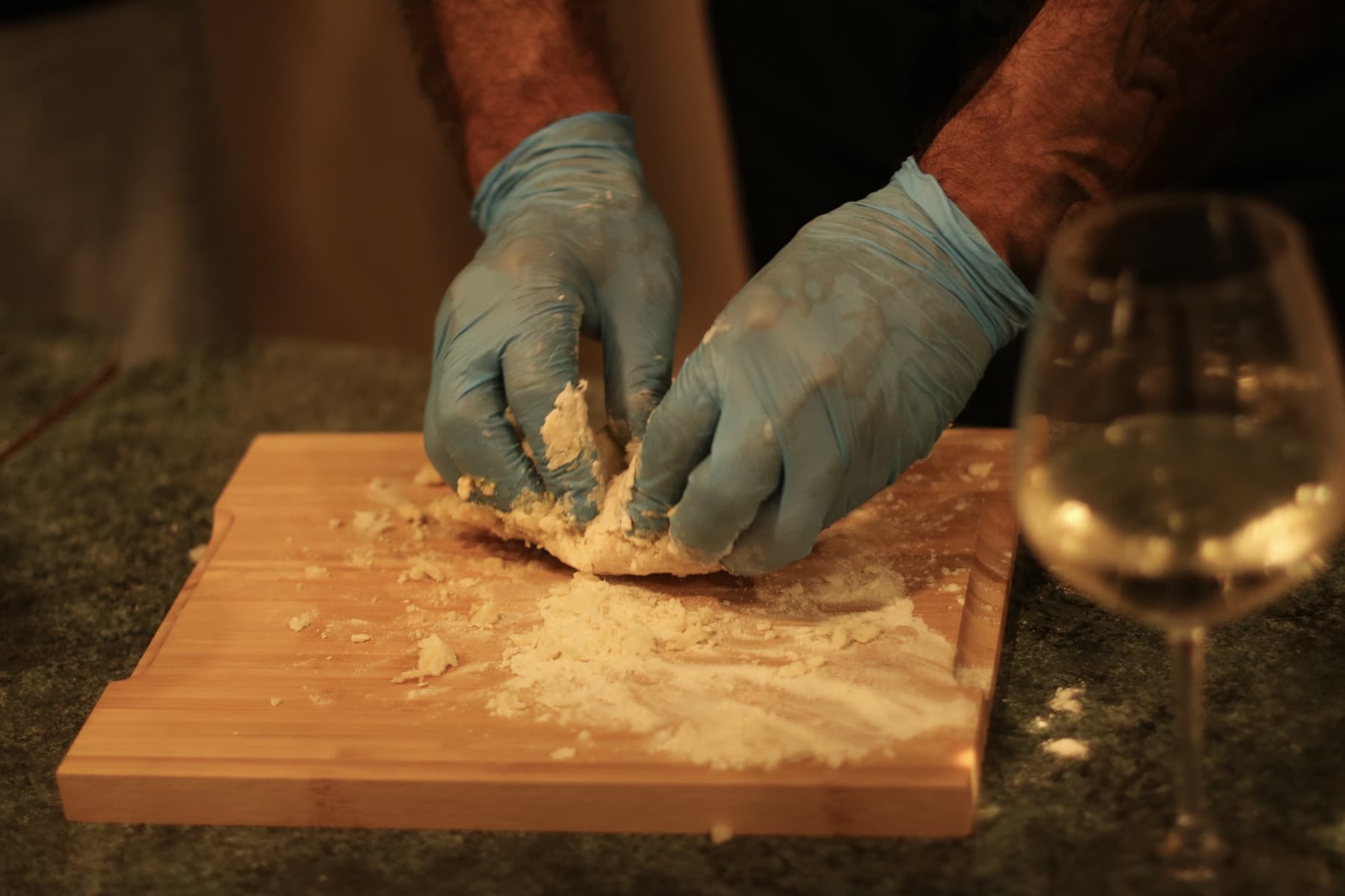 Gloved hands knead potato gnocchi dough on a wooden board next to a glass of white wine