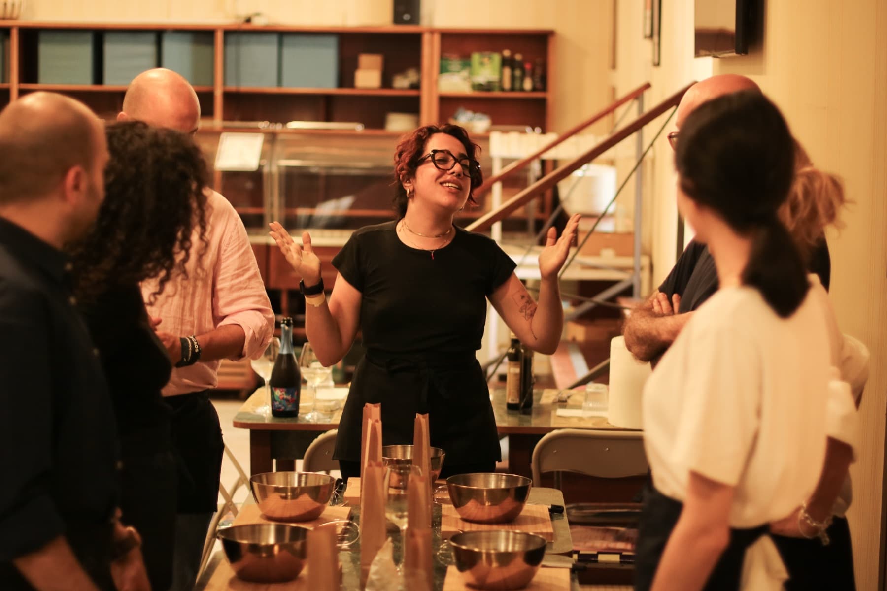 The host, arms spread, explains the next step to guests gathered around a table