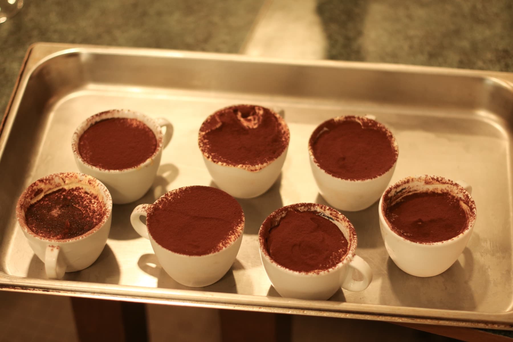 Seven homemade tiramisu served in white espresso cups, dusted with cocoa, lined up on a steel tray